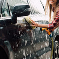 Person washing a black car with sponge, soap suds, and hose outdoors