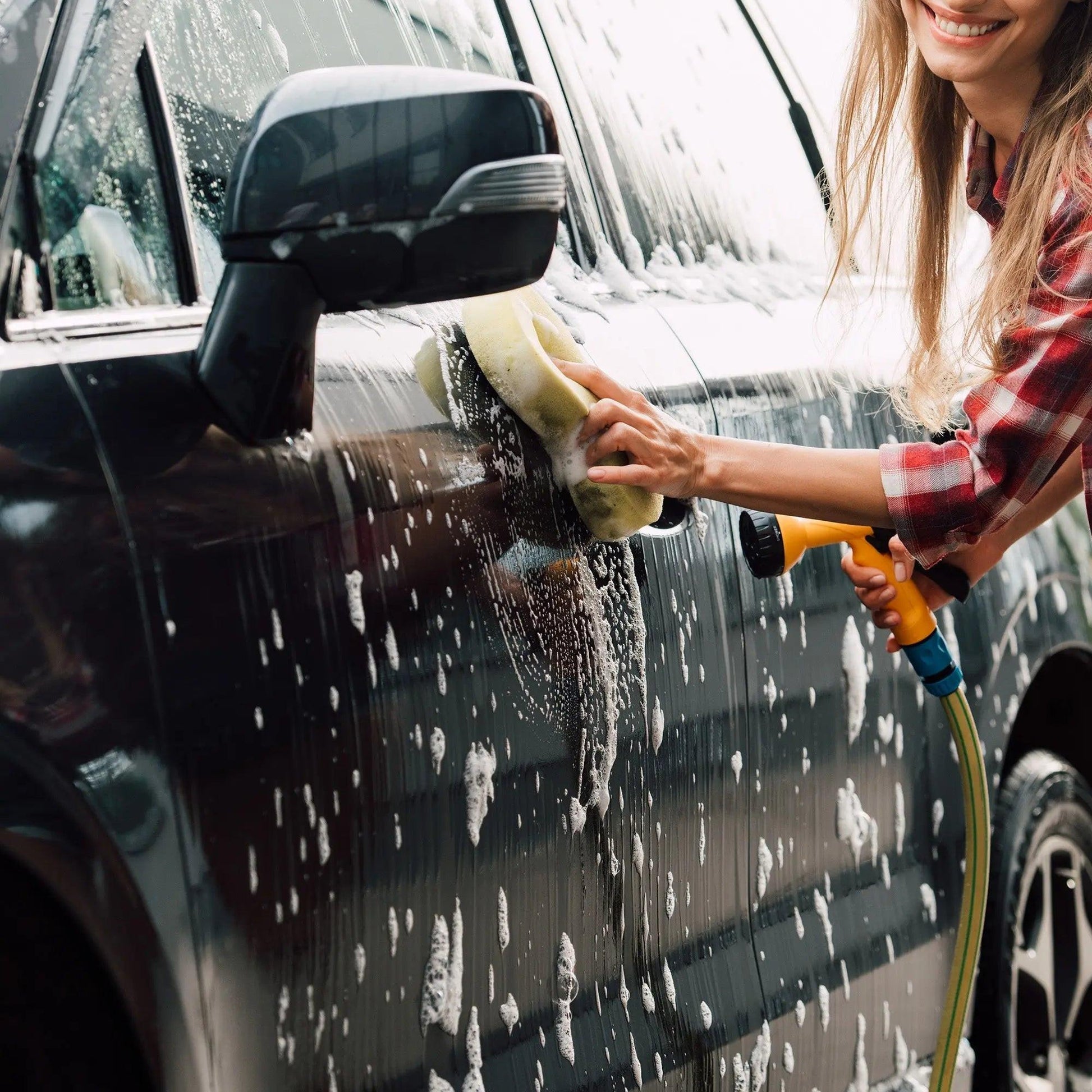 Person washing a black car with sponge, soap suds, and hose outdoors
