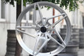 Silver multi-spoke alloy wheel for Audi, displayed outdoors with trees and buildings in background.