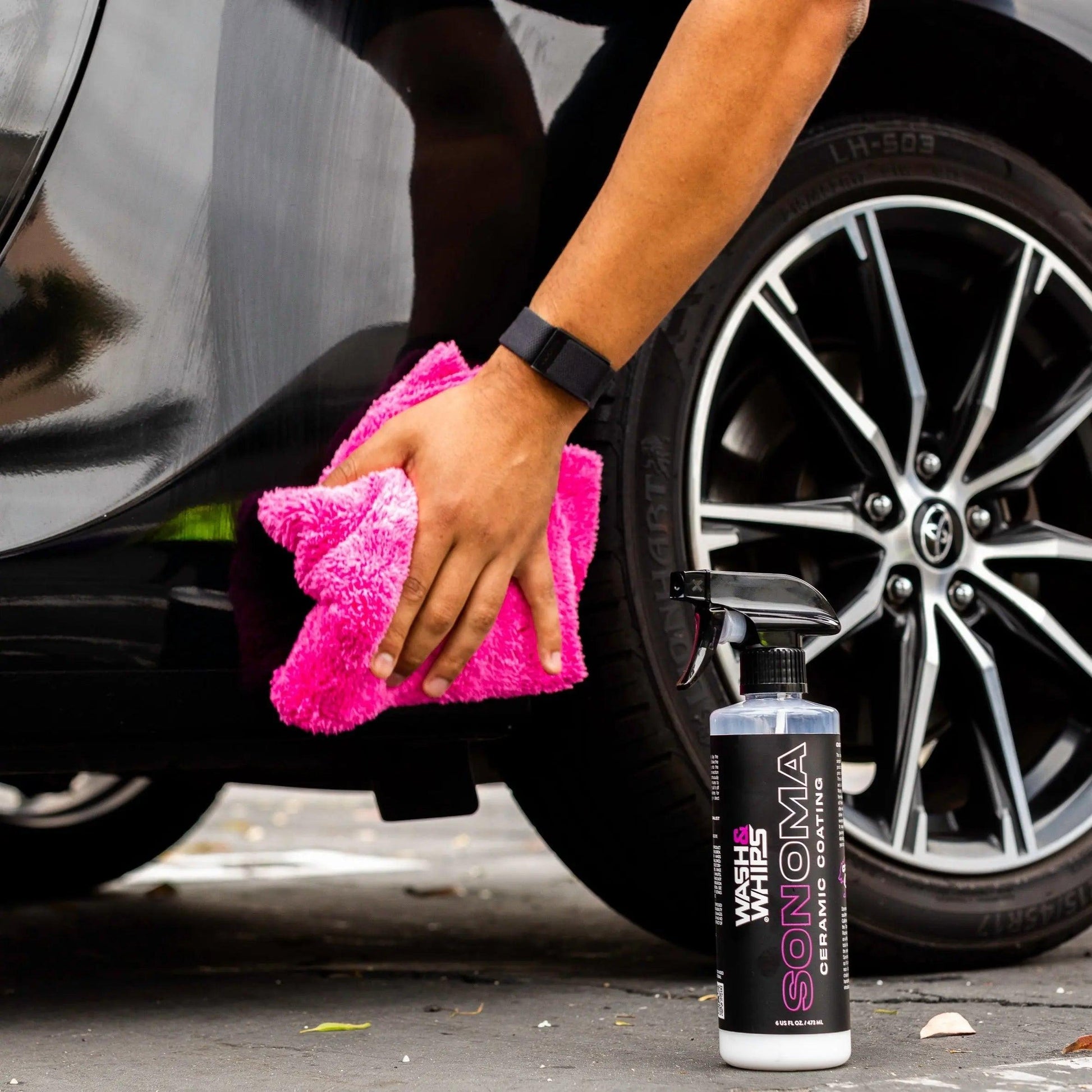Hand cleaning black car with pink microfiber towel next to ceramic coating spray bottle