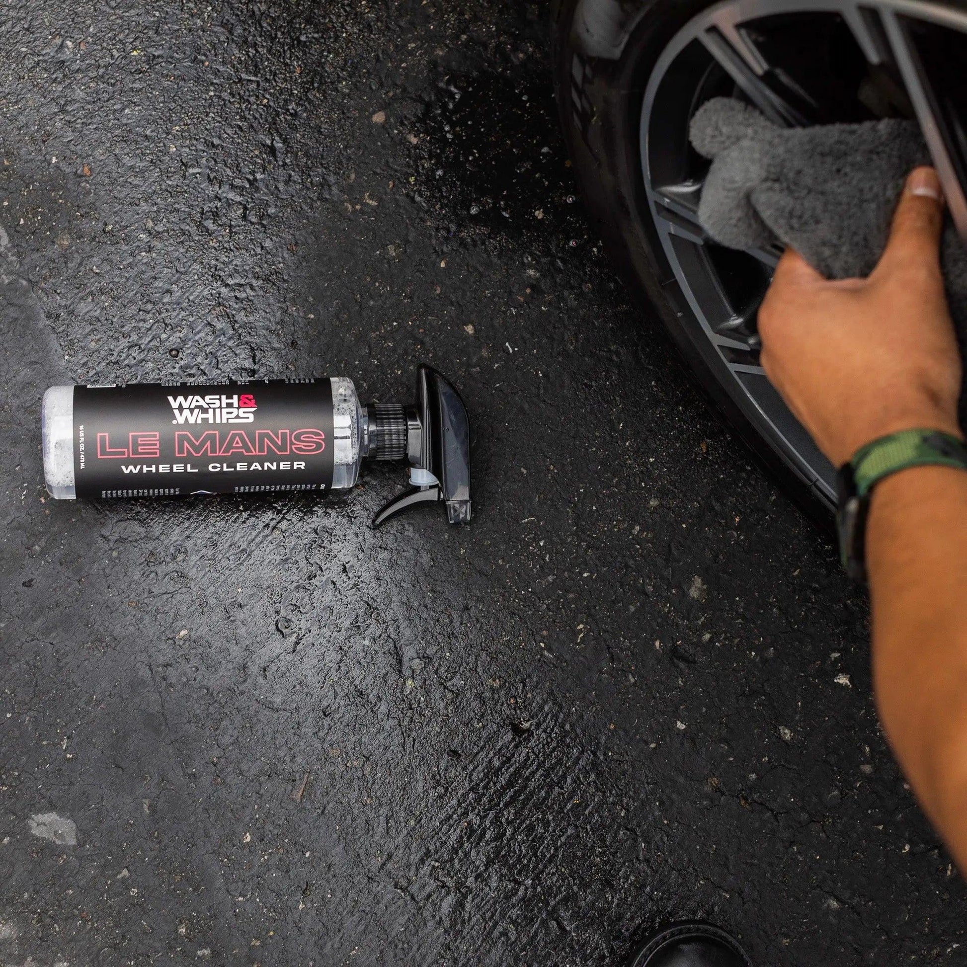 Person cleaning car wheel with Wash & Whips Le Mans wheel cleaner spray on wet pavement