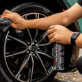 Person applying tire shine spray to a clean black car wheel with microfiber cloth.
