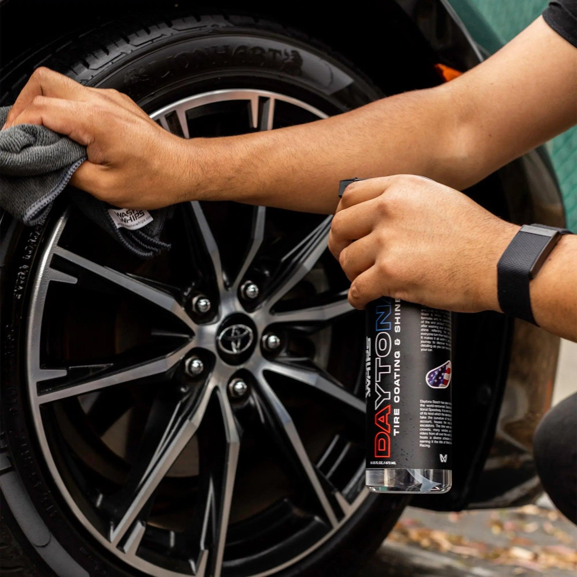 Person applying tire shine spray to a clean black car wheel with microfiber cloth.