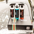 Car cleaning kit with wheel cleaner, car wash shampoo, and insect remover bottles on a foamy hood