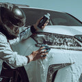 Person wearing racing suit and helmet washing white car headlight with soap and sponge